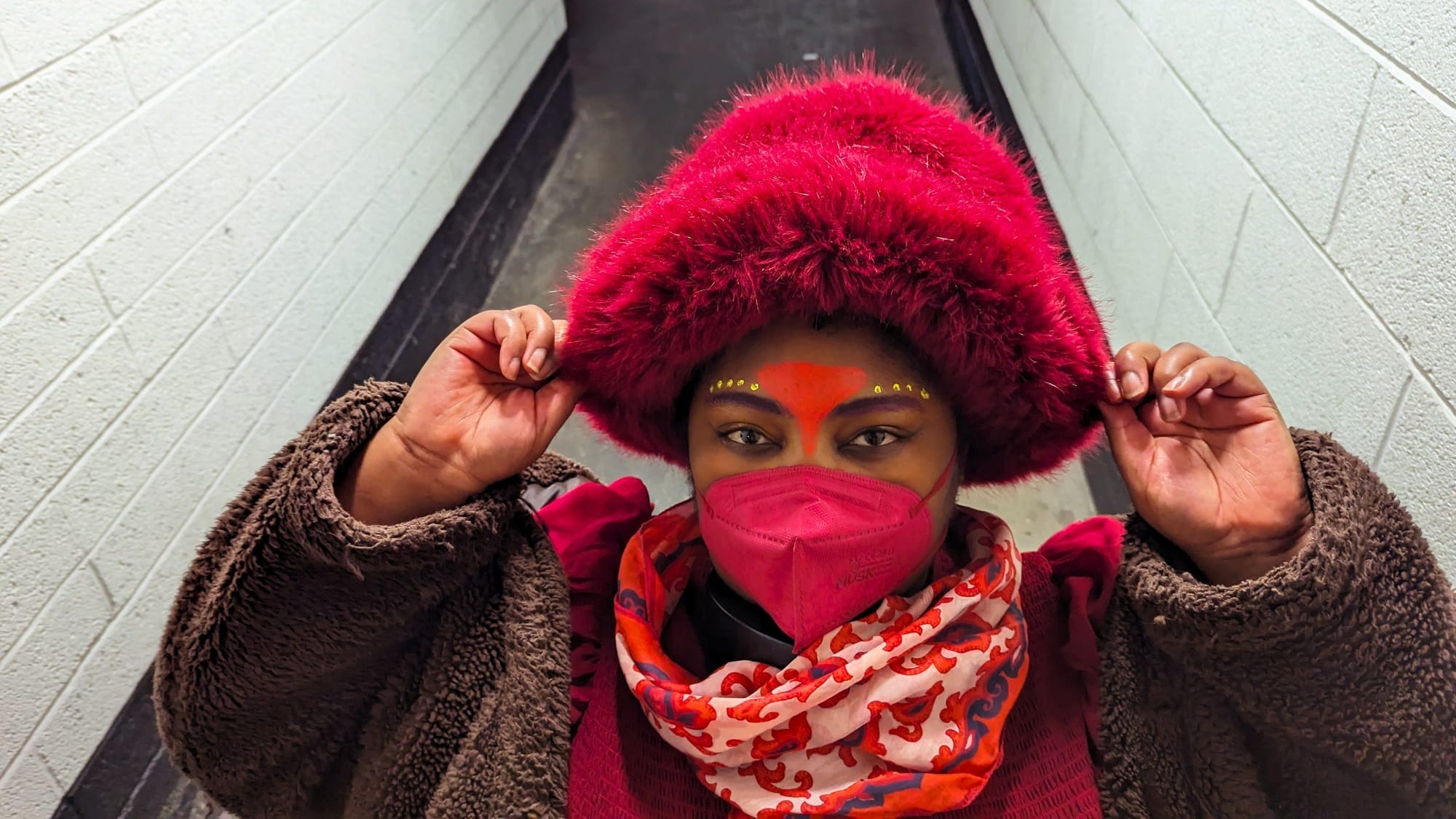 West African person in a hallway, wearing red and brown with a red kn95 mask