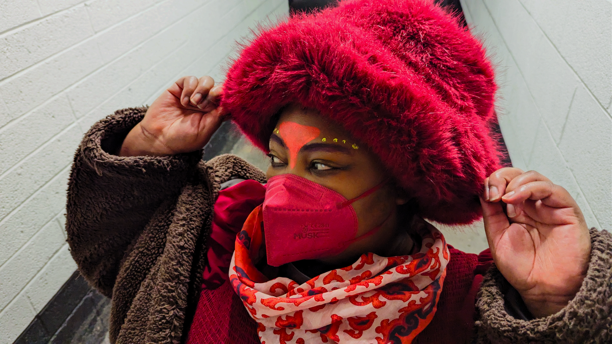 Love, Builder of Worlds is wearing a furry burgundy hat and n95 mask. They have colourful makeup on, red scarf and brown coat.