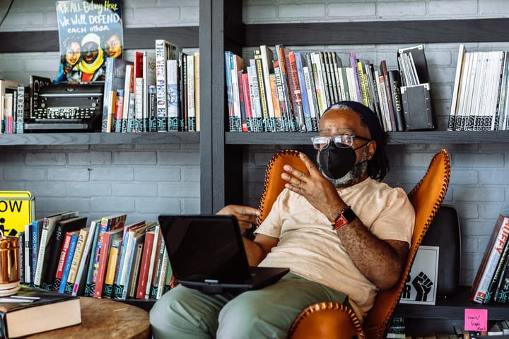 A Deaf Black man wearing glasses and a KN-95 mask leans back in a chair in a space with books behind