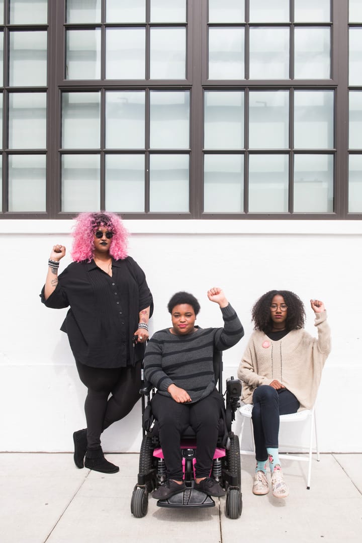 Three Black and disabled folx raise fists in front of a white wall.