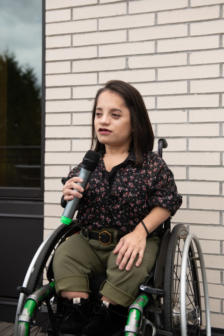 A South Asian person is in her wheelchair sitting in front of a brick wall, holding a microphone while giving a speech.