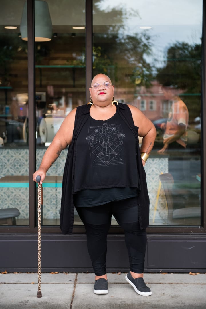 A Black non-binary person stands casually outside a cafe while leaning on their leopard print cane.